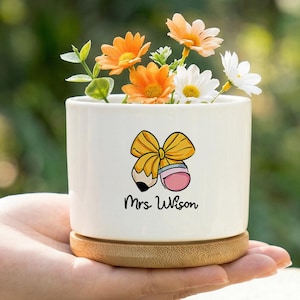 May include: A white ceramic planter with a bamboo base, filled with orange and white flowers. The planter features a cartoon bow and pencil design with the name "Mrs. Wilson" below. The planter is held in a hand.