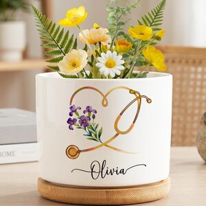 May include: A white cylindrical ceramic planter with a bamboo base, featuring a gold stethoscope graphic shaped into a heart, a small cluster of purple flowers, and the name Olivia printed in black script lettering on the front surface.