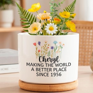 May include: A white ceramic cylindrical planter with a bamboo base, featuring a colorful wildflower graphic and black text that reads: Cheryl MAKING THE WORLD A BETTER PLACE SINCE 1956. The planter is filled with yellow and white artificial flowers.