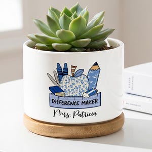 Puede incluir: Una maceta de cerámica blanca con una planta suculenta. La maceta tiene un diseño de manzana azul y blanco con las palabras "DIFFERENCE MAKER" y "Mrs. Patricia". Se apoya en un posavasos redondo de madera.