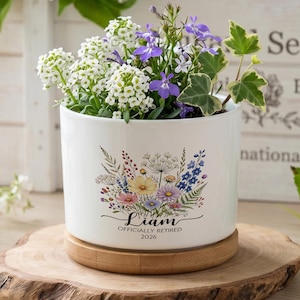 May include: A white ceramic planter with a floral design and the name "Liam" with "OFFICIALLY RETIRED 2026" printed below. The planter holds a variety of flowers and greenery and sits on a wooden base.