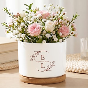 May include: A white ceramic flower pot with a wooden base, filled with pink and white roses and greenery. The pot has a decorative design with the letters "E" and "L" and the date "August 02, 2026". The pot is sitting on a wooden table.
