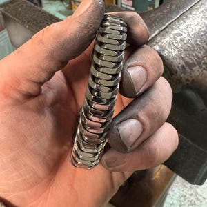 May include: A silver-colored, articulated bracelet with a unique, segmented design. The bracelet is held in a hand covered in dark residue, suggesting a workshop environment. The bracelet's links are rectangular and closely connected.