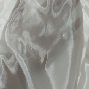 May include: A close-up of a sheer, white fabric. The fabric is slightly translucent, allowing a glimpse of the hand behind it. The material has a subtle sheen, with soft folds and highlights. The overall texture appears smooth and delicate.