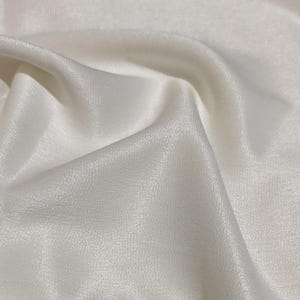 May include: Close-up of a flowing, off-white fabric with a subtle sheen. The material is draped, creating soft folds and shadows. The texture appears smooth and luxurious, suggesting a high-quality textile.