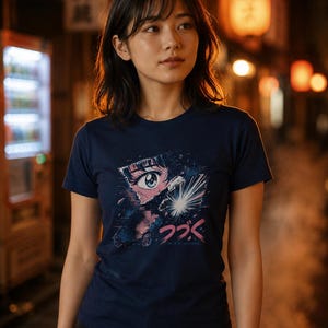 May include: A dark blue short-sleeved t-shirt featuring a graphic print of a stylized anime eye and a bright light burst. Below the graphic, the Japanese text reads "つづく" with the English text "TO BE CONTINUED" printed underneath in a smaller font.