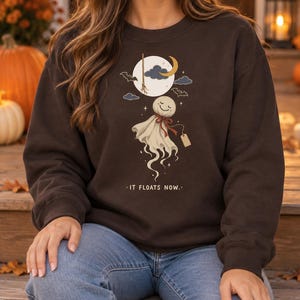 Puede incluir: Una sudadera de cuello redondo color marrón oscuro con un estampado gráfico de una figura fantasmal sonriente con un lazo rojo, una escoba, una luna creciente y murciélagos. El texto IT FLOATS NOW está impreso debajo de la ilustración.