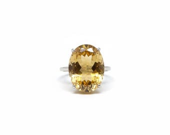 May include: A silver ring featuring a large, oval-cut, golden-yellow gemstone. The gemstone is faceted, catching the light and displaying a range of warm tones. The ring's band is a simple, thin silver band.