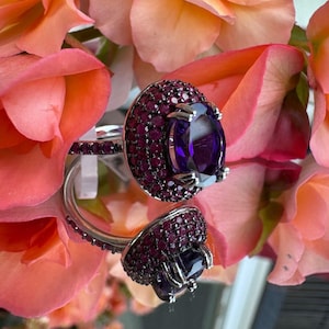 May include: A silver ring with a large, oval, deep purple gemstone at the center, surrounded by smaller, round, dark red gemstones. The ring is set against a backdrop of soft pink and orange roses.