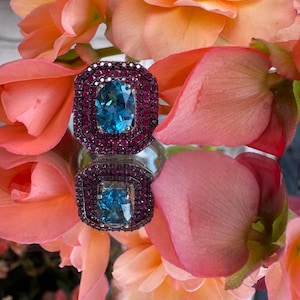 May include: A square-shaped ring with a large, oval, light blue gemstone at its center, surrounded by a border of smaller, dark red gemstones. The ring is set against a backdrop of peach-colored flowers.