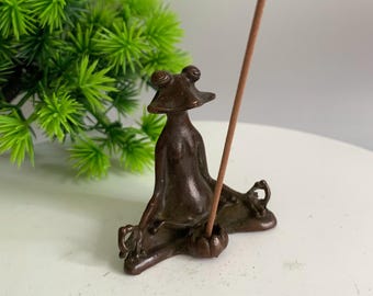 May include: A bronze frog figurine in a seated yoga pose, holding an incense stick. The frog has large eyes and a serene expression. The figurine is on a white surface with green foliage in the background.