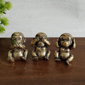 May include: Three bronze-colored monkey figurines in the "See no evil, hear no evil, speak no evil" pose. Each monkey is seated with its hands covering its eyes, ears, and mouth. The figurines are on a dark brown surface.