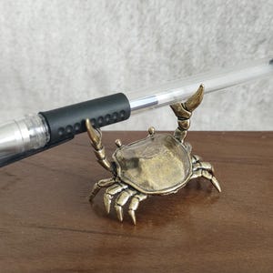 May include: A bronze crab pen holder with a black and clear pen resting in its claws. The crab is detailed with textured surfaces and is set on a wooden surface. The pen is approximately 15 cm long.