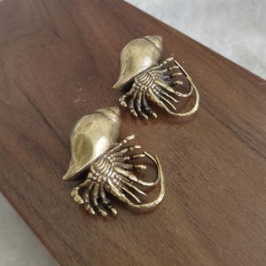 May include: Two brass-colored hermit crab sculptures. Each sculpture features a detailed crab body emerging from a spiral shell. The crab's legs and claws are intricately designed. The sculptures are placed on a dark wooden surface.