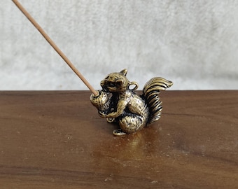 May include: A bronze-colored squirrel incense holder. The squirrel is holding an acorn and has a long, bushy tail. A thin, brown incense stick is inserted into the acorn. The holder is on a wooden surface.