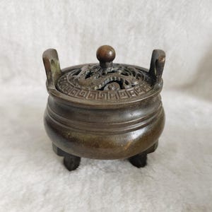 May include: A bronze-colored incense burner with a round body and three short legs. The lid features intricate cut-out designs and a small knob. Two handles are on either side of the burner.