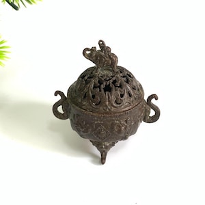 May include: A bronze-colored incense burner with an elephant figure on the lid. The burner has a round body with ornate cut-out patterns and curved handles. It stands on three small feet and is likely used for burning incense.