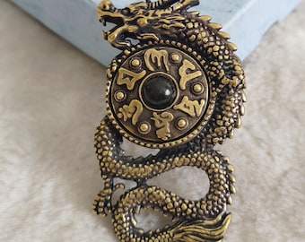 Puede incluir: Colgante de dragón de color bronce con detalles intrincados. El cuerpo del dragón rodea un emblema circular con una piedra negra y símbolos en relieve. El colgante está diseñado para colgar.