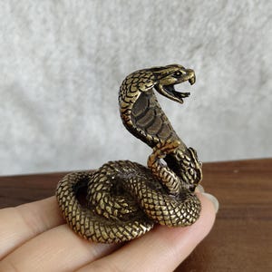 May include: A detailed brass cobra figurine, coiled and poised to strike. The snake's hood is raised, and its mouth is open, revealing its fangs. The sculpture has a textured, scale-like pattern and a vintage, antique finish.
