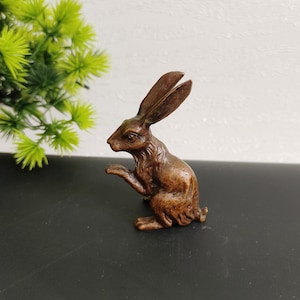 Op de afbeelding: Een bronzen konijnenbeeldje met lange oren en een opgeheven poot. Het konijn zit in een zittende positie, met gedetailleerde vachttextuur. Het beeldje staat op een zwart oppervlak met een groene plant op de achtergrond.