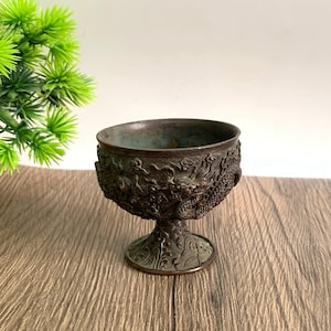 May include: A bronze-colored, ornate goblet with a detailed dragon design wrapping around the bowl. The goblet has a pedestal base and is set on a wooden surface. The cup is likely used for decorative purposes.