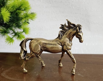 May include: A bronze-colored horse figurine in a dynamic pose, with a flowing mane and tail. The sculpture is detailed, capturing the horse's musculature and elegant form. The figurine is displayed on a wooden surface, with a green plant in the background.
