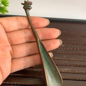 May include: A bronze spoon featuring a dragon head on the handle. The spoon has a long, thin handle and a wide, shallow bowl with a green patina. The spoon is held in a hand.