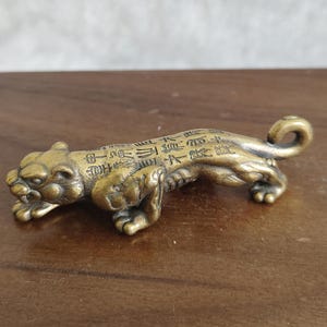 May include: A small, bronze-coloured tiger figurine. The tiger is in a crouching position with its head slightly raised. The body of the tiger is covered in engraved characters. The figurine is resting on a wooden surface.