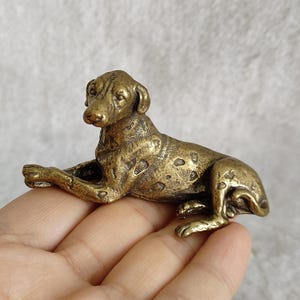 Pode incluir: Pequena estatueta de cão em bronze, em posição de repouso. O cão tem uma pelagem manchada e está deitado com as patas dianteiras estendidas. A estatueta é provavelmente feita de metal e tem uma superfície texturizada e detalhada.
