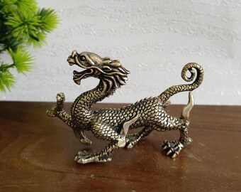 Puede incluir: Una detallada figura de dragón de color bronce con un cuerpo texturizado y escamoso y rasgos faciales intrincados. El dragón está posado en una postura dinámica, con la cola enroscada y la cabeza levantada. La figura se exhibe sobre una superficie marrón oscuro.