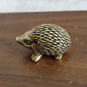 Peut inclure: Petite figurine de hérisson en laiton doré. Le corps est texturé, avec de petits yeux et quatre petites pattes. La figurine est de couleur dorée et repose sur une surface en bois.