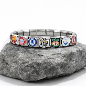 Puede incluir: Una pulsera de abalorios de acero inoxidable con múltiples abalorios de esmalte de colores que representan logotipos de equipos de fútbol. La pulsera se muestra sobre una piedra gris sobre un fondo blanco. Los logotipos incluyen Chelsea, Juventus y Liverpool.