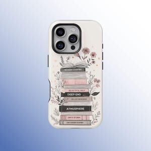 Op de afbeelding: Witte telefoonhoes met een ontwerp van gestapelde boeken, waaronder titels als "Broken Country" en "Deep End". De hoes is versierd met roze en witte bloemmotieven. De cameramodule van de telefoon is zwart.