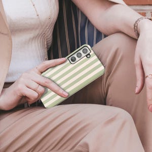 May include: A phone case with a green and cream striped pattern. The case has a rectangular shape with rounded corners and a camera cutout. The case is held by a person wearing rings and a bracelet.