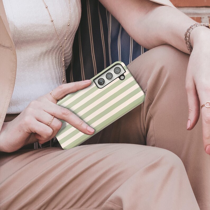 May include: A phone case with a green and cream striped pattern. The case has a rectangular shape with rounded corners and a camera cutout. The case is held by a person wearing rings and a bracelet.
