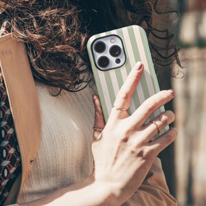 May include: A mint green and white striped phone case. The case is held in a hand with gold rings. The phone case has a black camera lens area. The background is blurred, suggesting an outdoor setting.