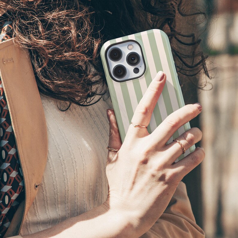 May include: A mint green and white striped phone case. The case is held in a hand with gold rings. The phone case has a black camera lens area. The background is blurred, suggesting an outdoor setting.