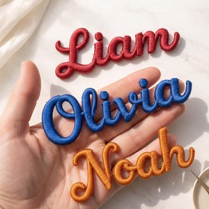 Puede incluir: Parches de nombres bordados en letras rojas, azules y naranjas. Se muestran los nombres "Liam", "Olivia" y "Noah". Un plato pequeño con útiles de costura, incluyendo tijeras, botones e hilo, está en el fondo.