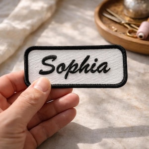 May include: A rectangular white patch with the name "Sophia" embroidered in black cursive lettering. The patch has a black border and is held in a hand. The background includes a wooden tray with sewing tools.