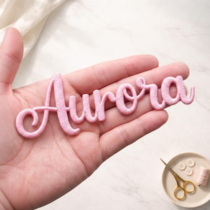 May include: A pink embroidered patch in a cursive font that spells "Aurora". The patch is held in a hand, with a small plate of sewing supplies in the background, including scissors, buttons, and thread.