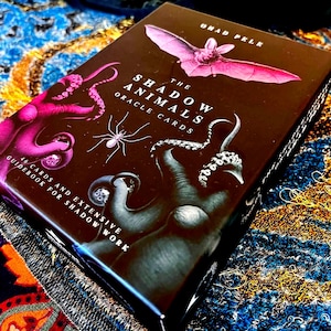 May include: A rectangular box for The Shadow Animals Oracle Cards by Ohad Pele. The black cover features a pink bat, a white spider, and dark octopus tentacles. Text includes 46 cards and extensive guidebook for shadow work in white font.