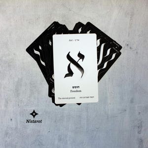 Könnte beinhalten: Ein Deck aus schwarz-weißen Tarotkarten. Die zentrale Karte zeigt ein schwarzes Alef-Symbol und das Wort "Freedom". Die Karten sind auf einer grauen Oberfläche angeordnet, mit dem Markennamen "Nistarot" sichtbar.