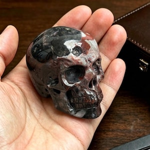 Puede incluir: Una talla de calavera en piedra pulida con una paleta de colores moteados en negro profundo, rojo oscuro e inclusiones minerales blancas. La escultura muestra rasgos anatómicos realistas, incluyendo cuencas oculares huecas y dientes.