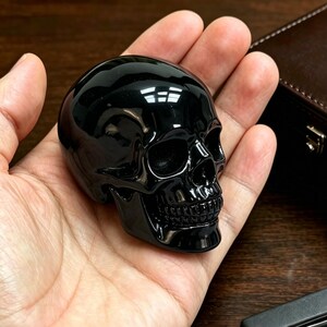 Könnte beinhalten: Eine glänzende, schwarze Schädel-Figur mit detaillierten Gesichtszügen. Der Schädel wird in einer Hand gehalten, was seine polierte Oberfläche und das komplizierte Design der Zähne und Augenhöhlen zeigt. Die dunkle Farbe und glatte Textur verleihen ihm ein auffälliges Aussehen.