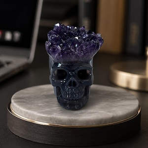Puede incluir: Una escultura de cráneo de superficie lisa en color azul oscuro, con un grupo de cristales de amatista de color púrpura intenso que emergen de la parte superior. El cráneo presenta rasgos faciales realistas, incluyendo cuencas oculares.