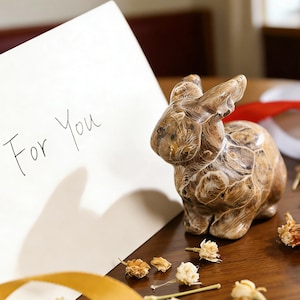 May include: A brown and beige rabbit figurine with a textured surface sits on a wooden table. A white card with the words "For You" is visible. Dried flowers and a gold ribbon are also present.