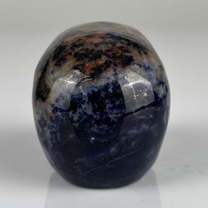 May include: A polished, smooth stone carving shaped like a human skull. The mineral features a mottled pattern of deep navy blue, dark black, and earthy brown patches, with a glossy, reflective surface that highlights the natural stone texture.