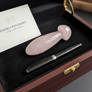 Puede incluir: Una herramienta de masaje de cuarzo rosa y un bolígrafo negro se exhiben dentro de una caja de madera con un forro de terciopelo negro. También es visible una tarjeta con el texto "ENERGY PATHWAYS: A Guide to Self-Connection". La caja está abierta, revelando el contenido.