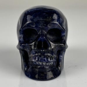 May include: A polished, dark blue stone carving shaped like a human skull. The surface features deep navy and indigo hues with subtle, lighter blue marbling throughout. The carving displays detailed eye sockets, a nasal cavity, and defined teeth.