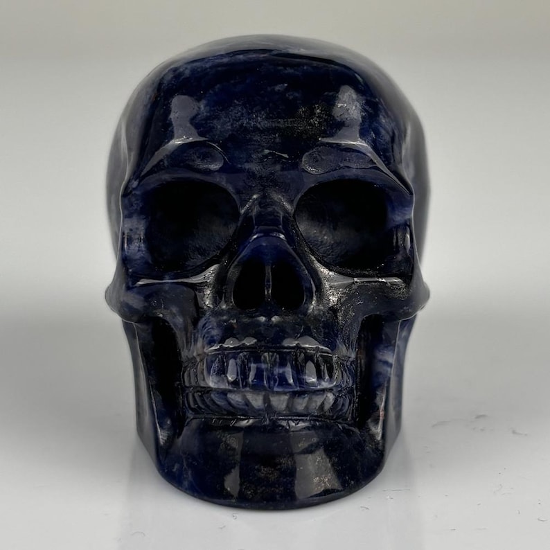 May include: A polished, dark blue stone carving shaped like a human skull. The surface features deep navy and indigo hues with subtle, lighter blue marbling throughout. The carving displays detailed eye sockets, a nasal cavity, and defined teeth.
